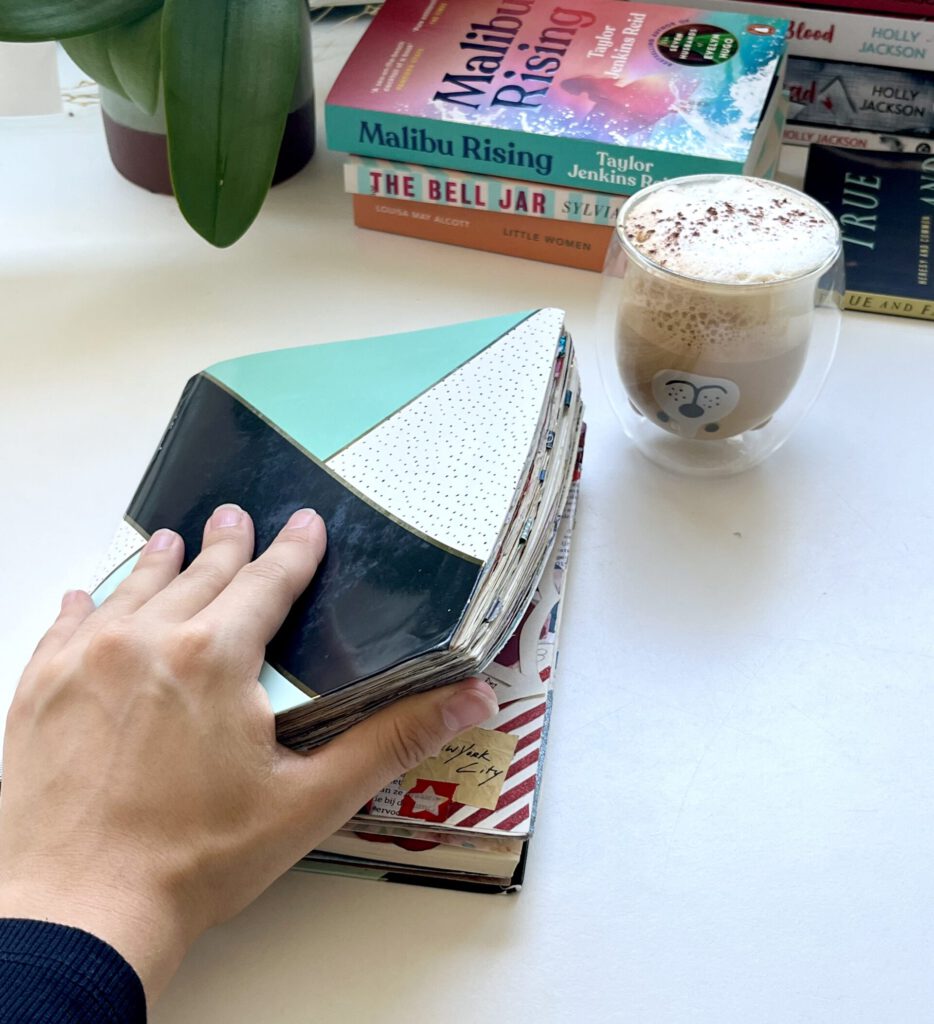 A bullet journal laying on a desk and being opened by a hand, with coffee next to it. In the background there are stacks of books and plants